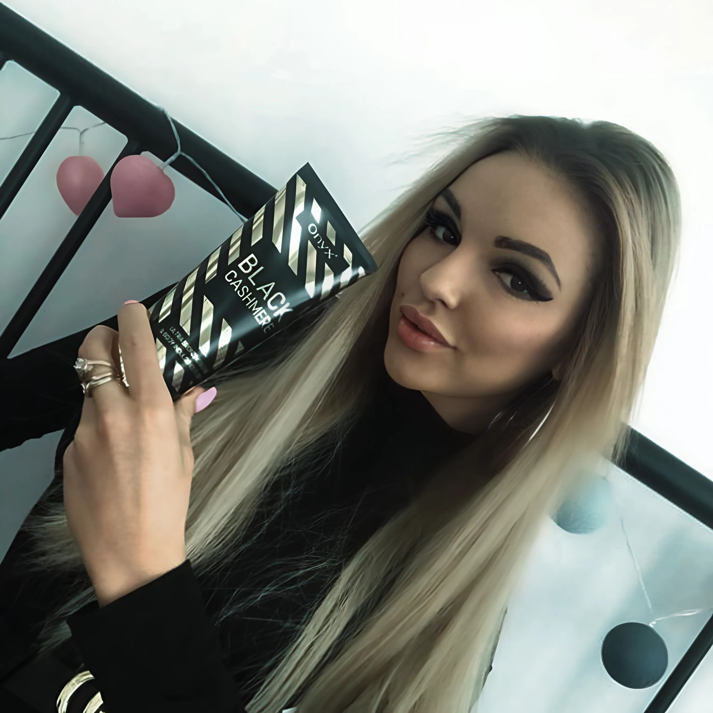 Blonde woman with sleek hair holding Onyx Black Cashmere tanning cream and smiling subtly at the camera