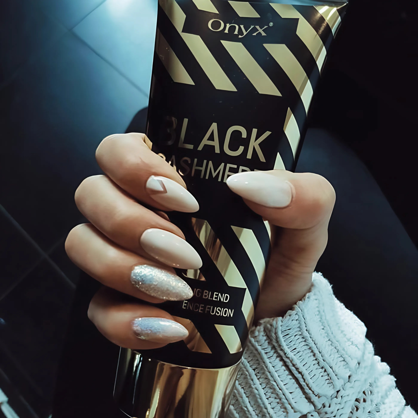 Hand with pale, glittery nails holding Onyx Black Cashmere tanning cream, creating a refined contrast with the dark background