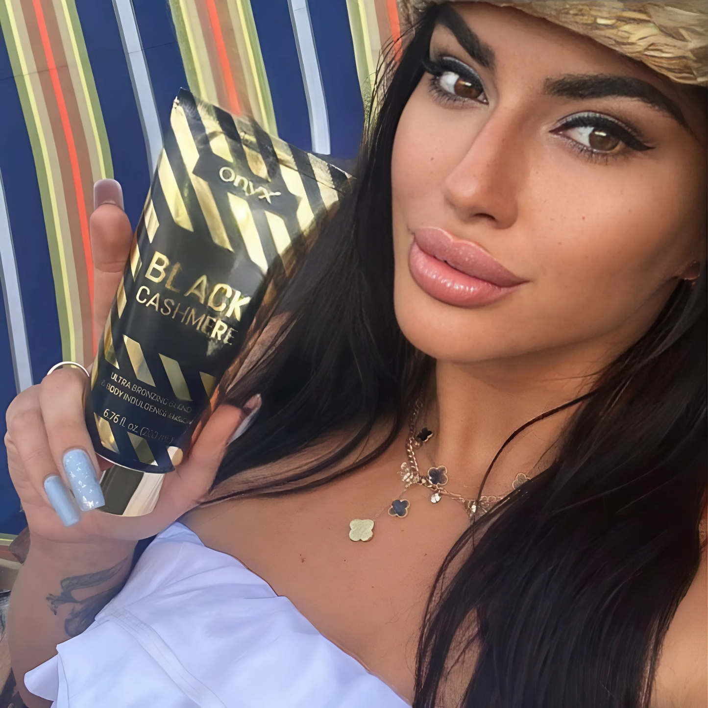 Woman with dark hair holding Onyx Black Cashmere tanning cream, the golden packaging gleaming in the sunlight