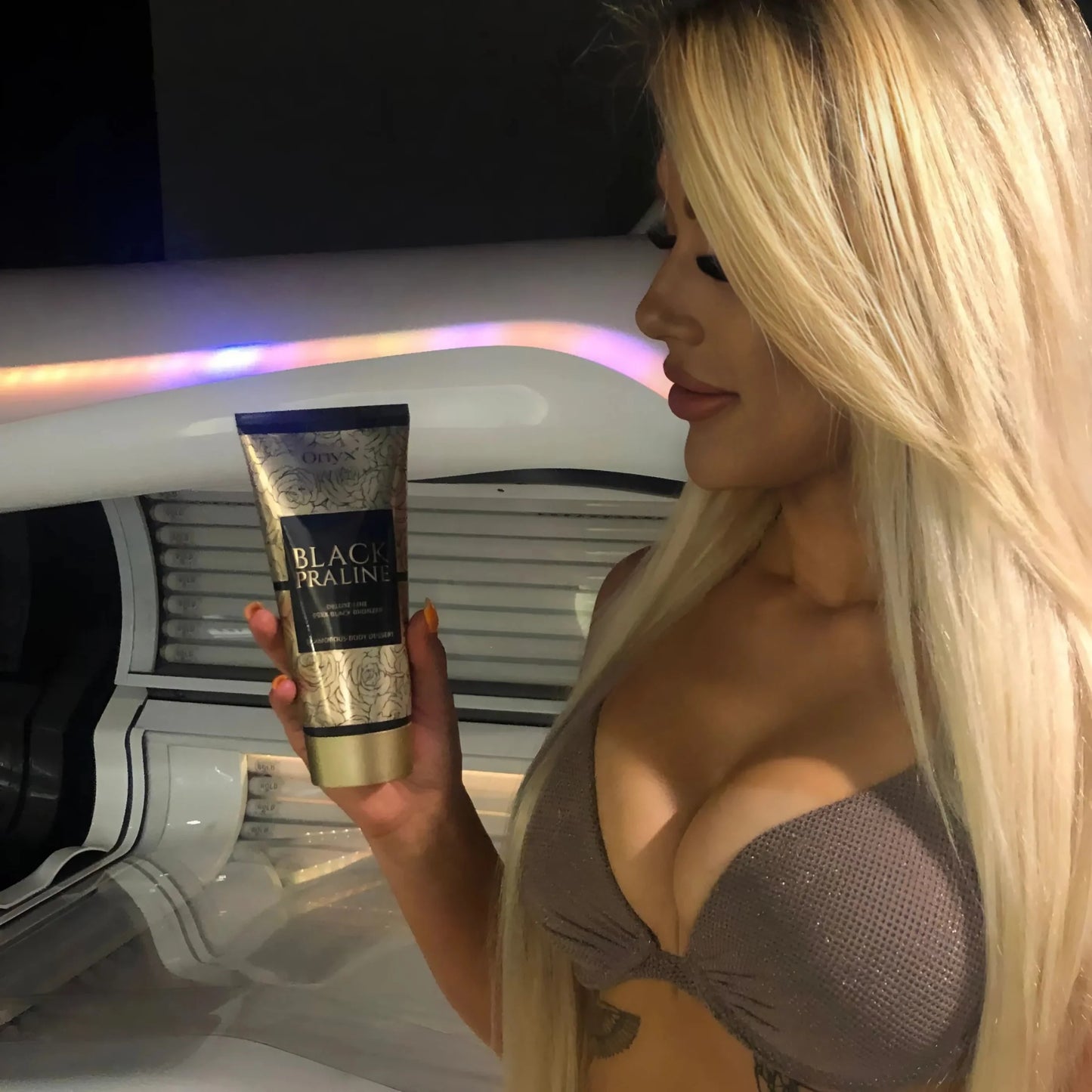 Blonde woman in a sunbed holding Onyx Black Praline tanning cream and admiring the luxurious golden tube under warm light