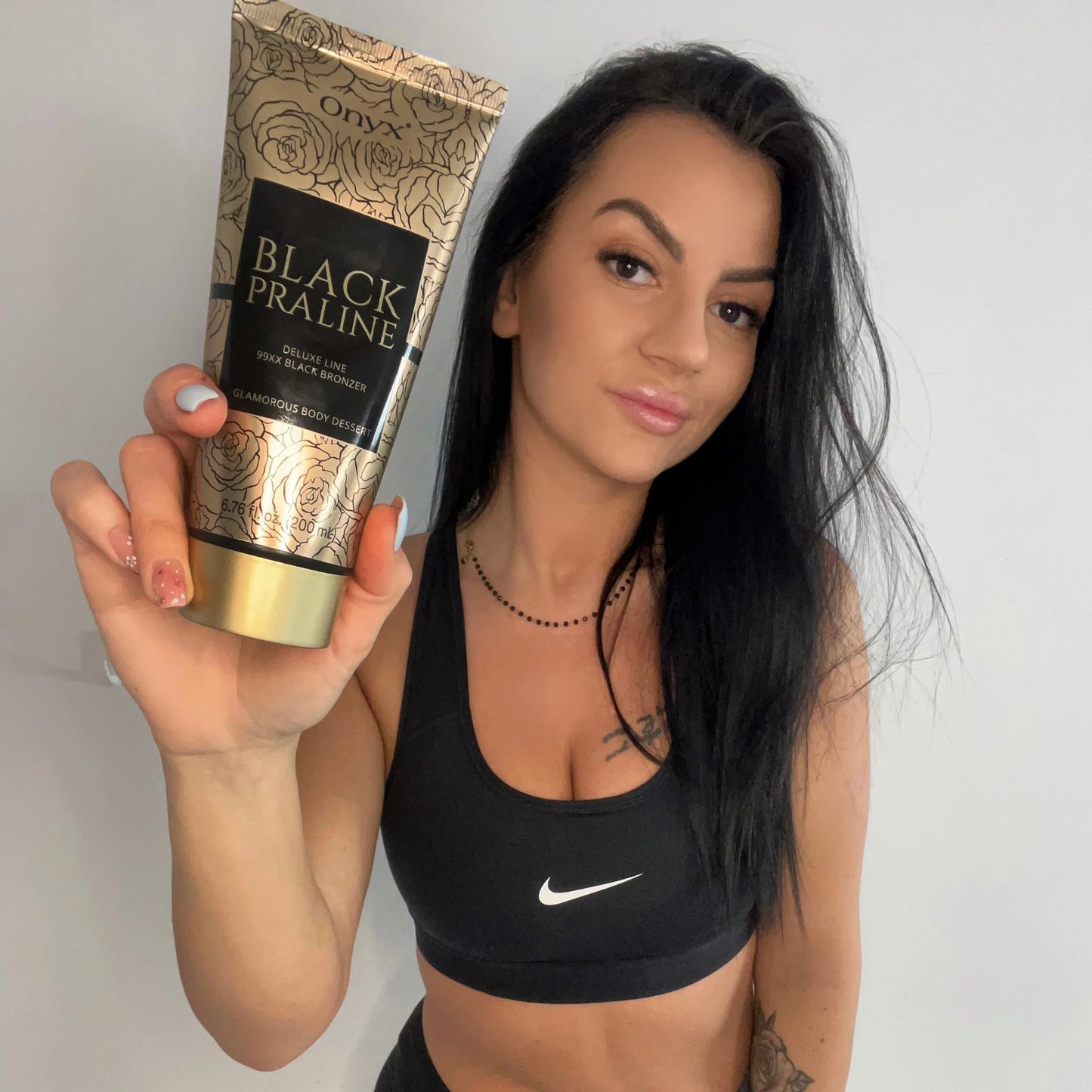 Woman with dark hair wearing a black sports bra presenting Onyx Black Praline tanning cream against a neutral background