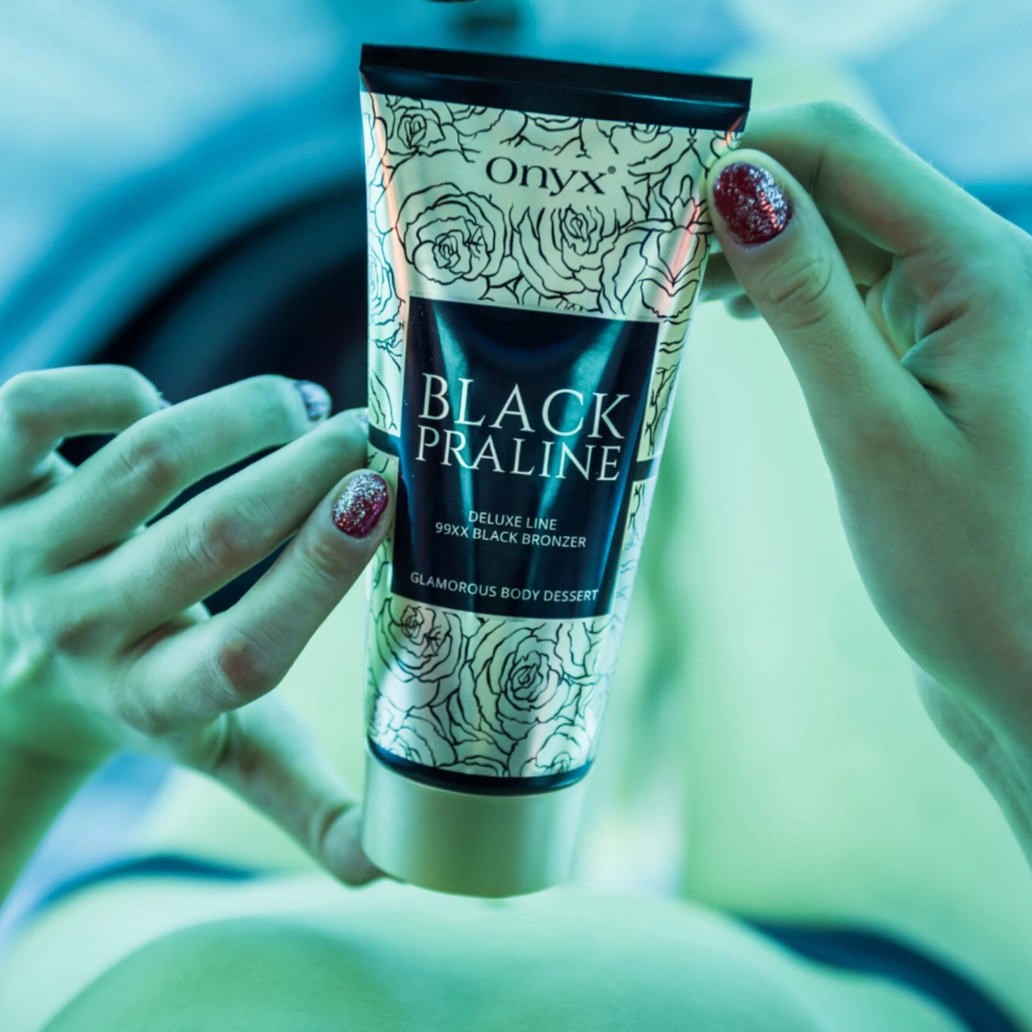 Onyx Black Praline tanning cream held by hands with glitter polish under soft blue sunbed light