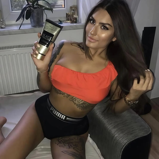 Dark-haired woman with tattoos sitting relaxed on a sofa, holding Onyx Black Praline tanning cream and playing with her hair