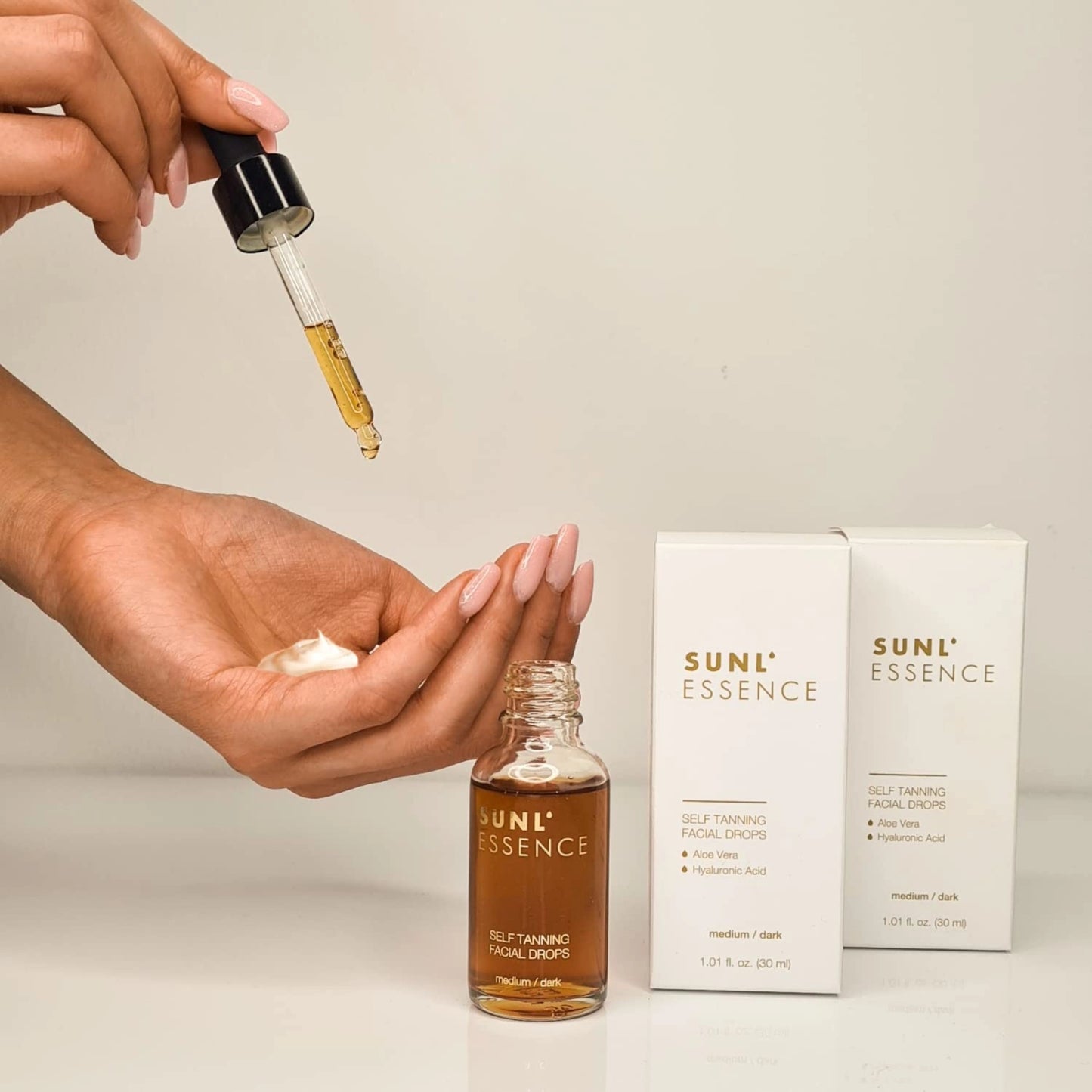 Hand applying self-tanning drops into the palm with a pipette next to two product boxes on a beige surface