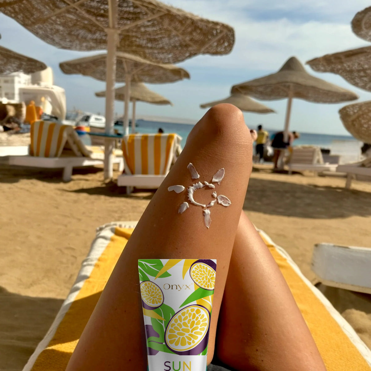 Tanned woman relaxing on a beach lounger with Onyx Suncolada Accelerator sunbed cream beside her leg and a sun drawn on her skin, showing smooth bronzed tan under parasols by the sea.