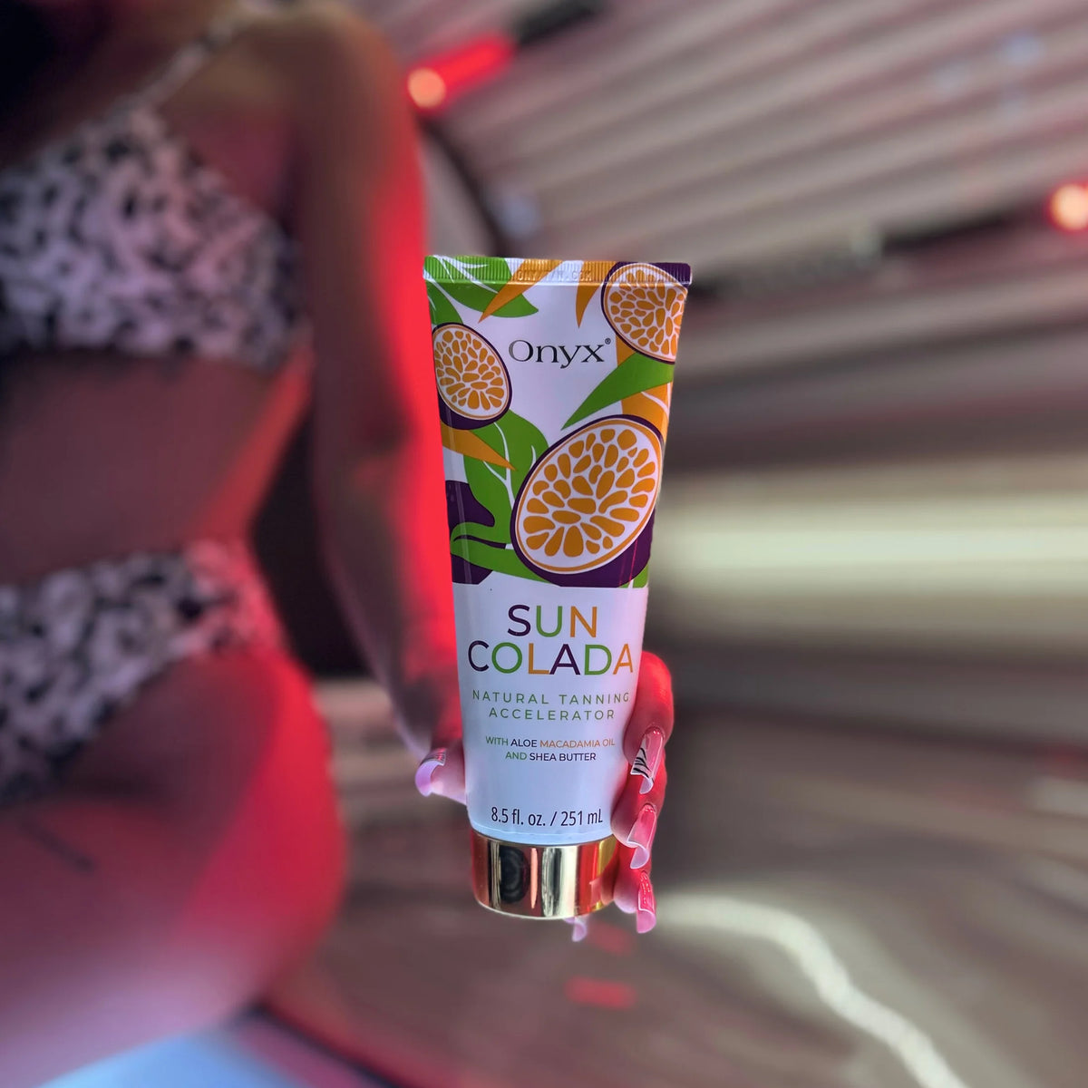 Close-up of an Onyx Suncolada Natural Tanning Accelerator cream tube held in front of a sunbed, with a model blurred in the background.