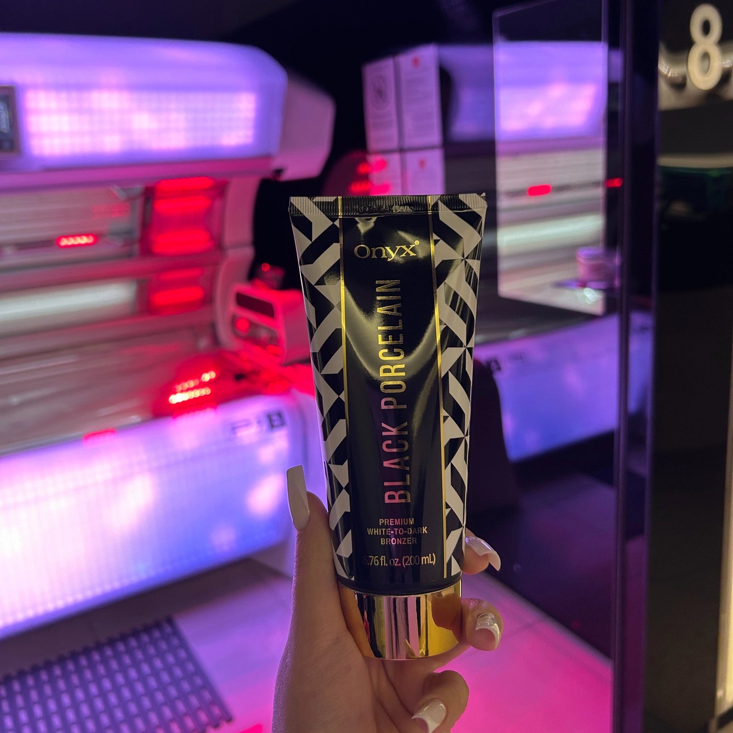 Hand with long white nails holding Onyx Black Porcelain cream in front of red-light tanning bed inside salon