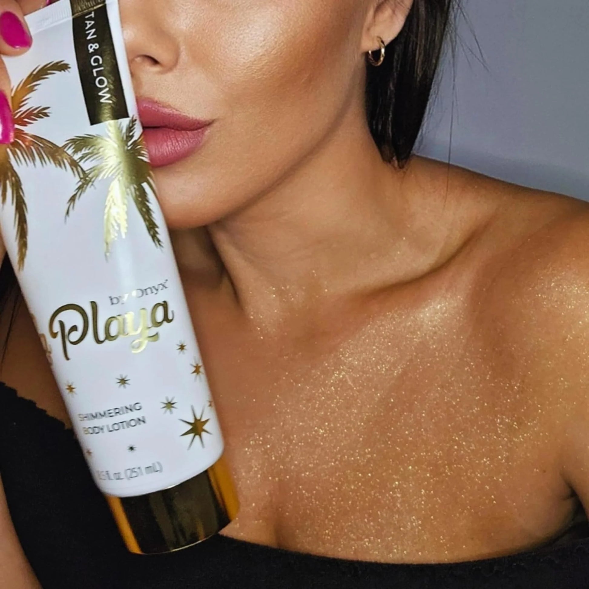 Close-up of a woman’s chest and shoulder glowing with shimmer as she holds a tube of shimmering body cream near her face