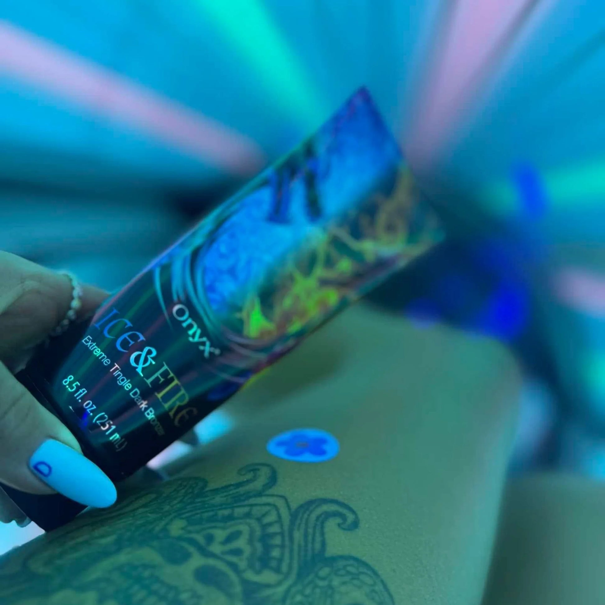Hand holding Onyx Ice & Fire tanning lotion during blue-lit sunbed session, tattoo visible on the leg.
