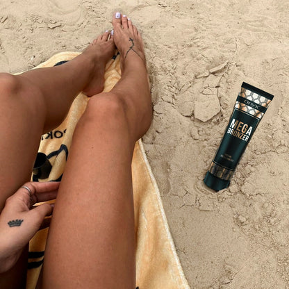 Close-up of a woman’s tanned legs resting on a sandy beach with Onyx Mega Bronzer cream lying nearby
