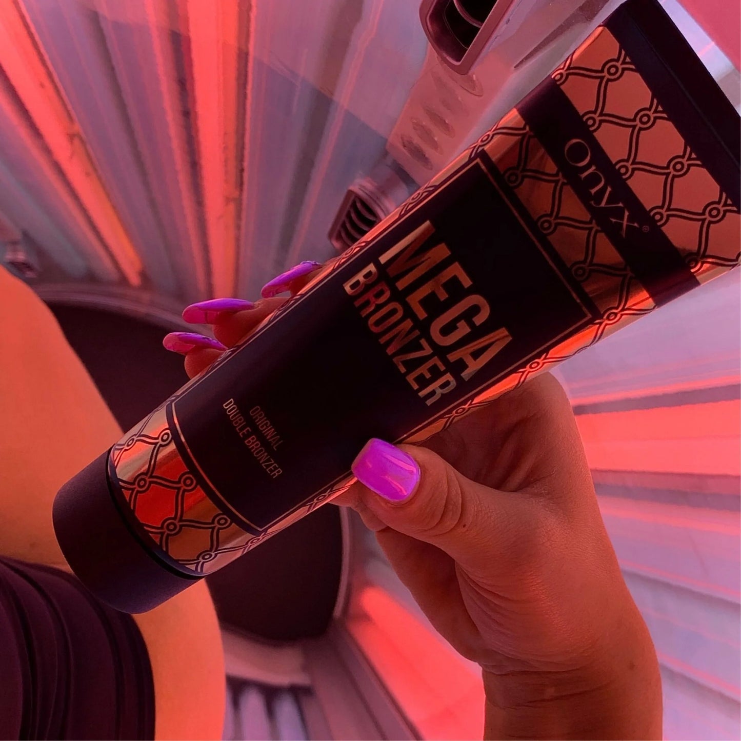 Hand with purple nails holding Onyx Mega Bronzer lotion under red lights inside a tanning bed