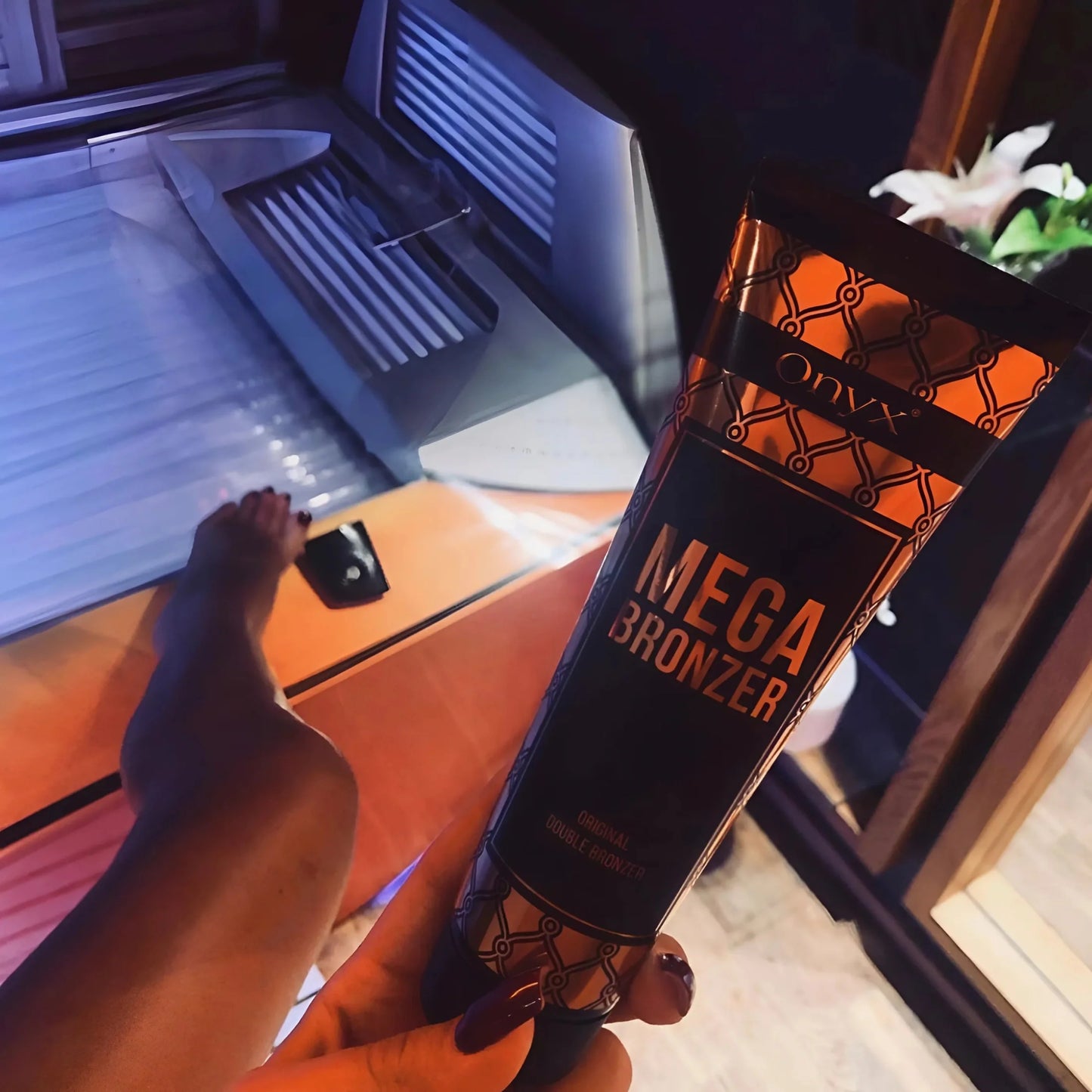 A woman holding Onyx Mega Bronzer tanning cream beside a sunbed, with focus on her tanned leg and warm ambient lighting