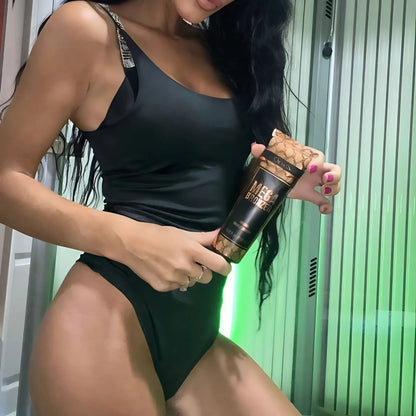 A woman in a black swimsuit holding Onyx Mega Bronzer tanning cream inside a sunbed room, showing her bronzed skin under soft green light