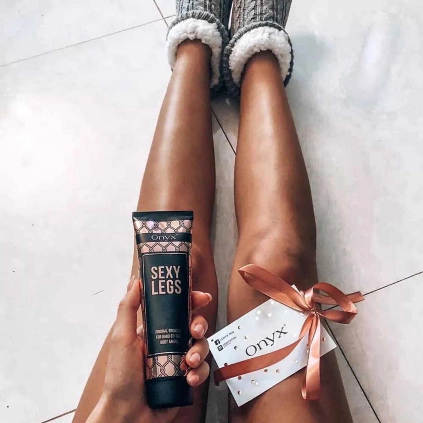 Woman sitting on the floor holding Onyx Sexy Legs cream beside her bronzed legs and a ribboned gift card