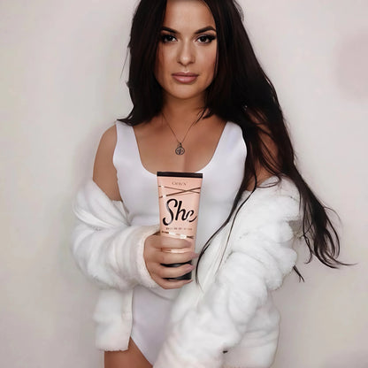 Woman in white bodysuit and soft fur jacket holding Onyx She bronzing cream while standing against a neutral wall