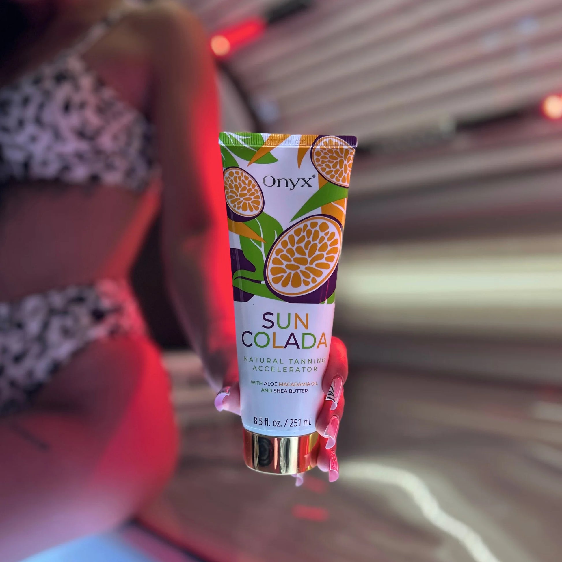 Close-up of an Onyx Suncolada Natural Tanning Accelerator cream tube held in front of a sunbed, with a model blurred in the background.