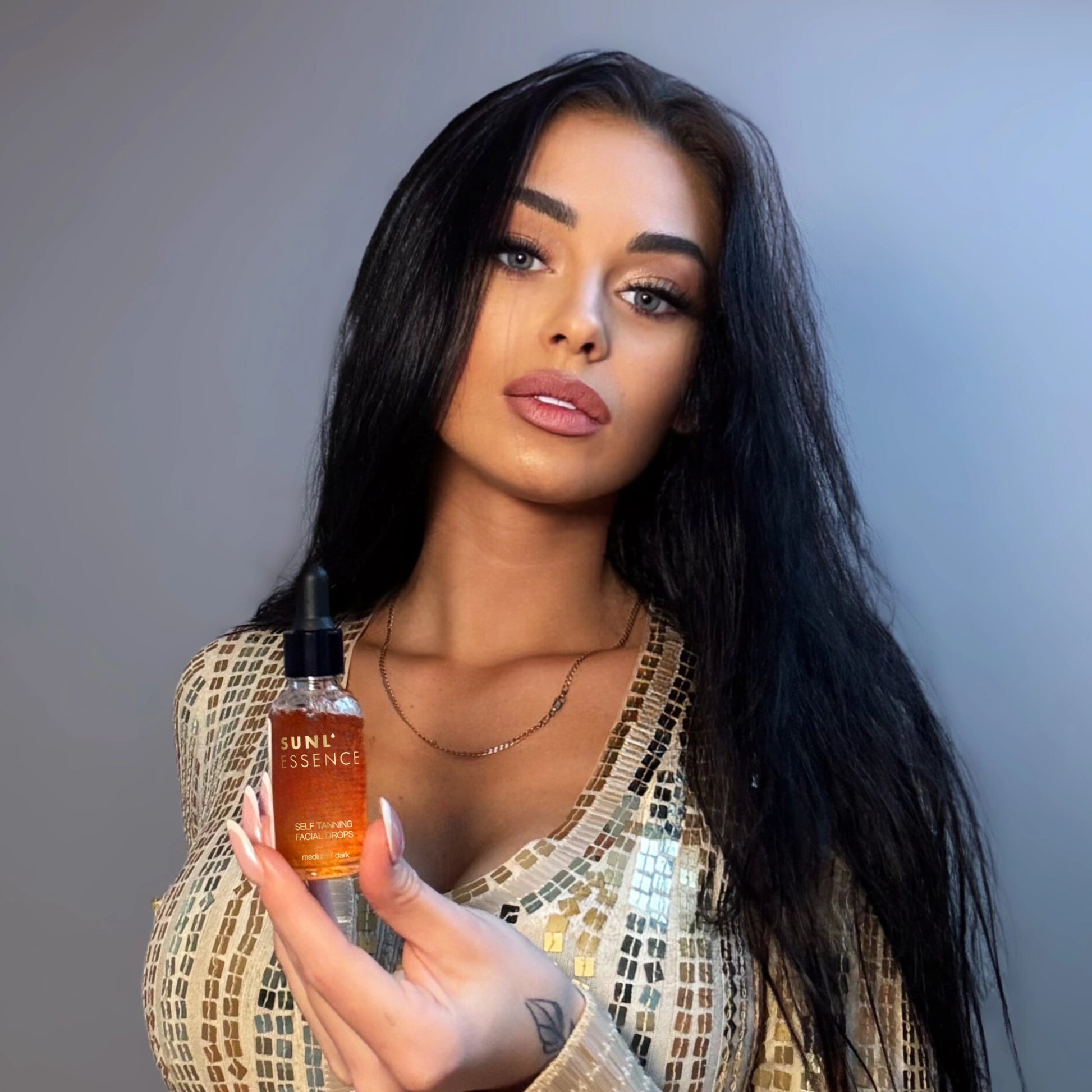 Portrait of a woman with long dark hair in a sparkly gold outfit holding a small bottle of self-tanning facial drops