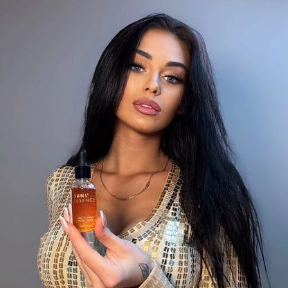Portrait of a woman with long dark hair in a sparkly gold outfit holding a small bottle of self-tanning facial drops