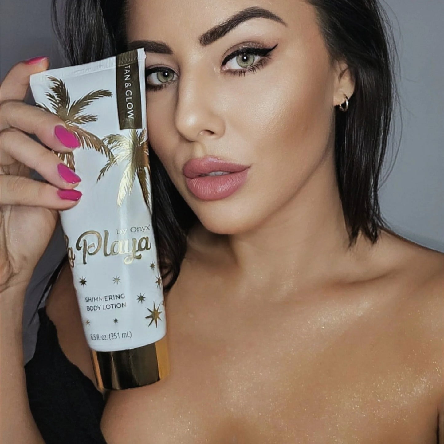Woman with dark hair holding a tube of shimmering body cream close to her face, showing glowing skin on her shoulders