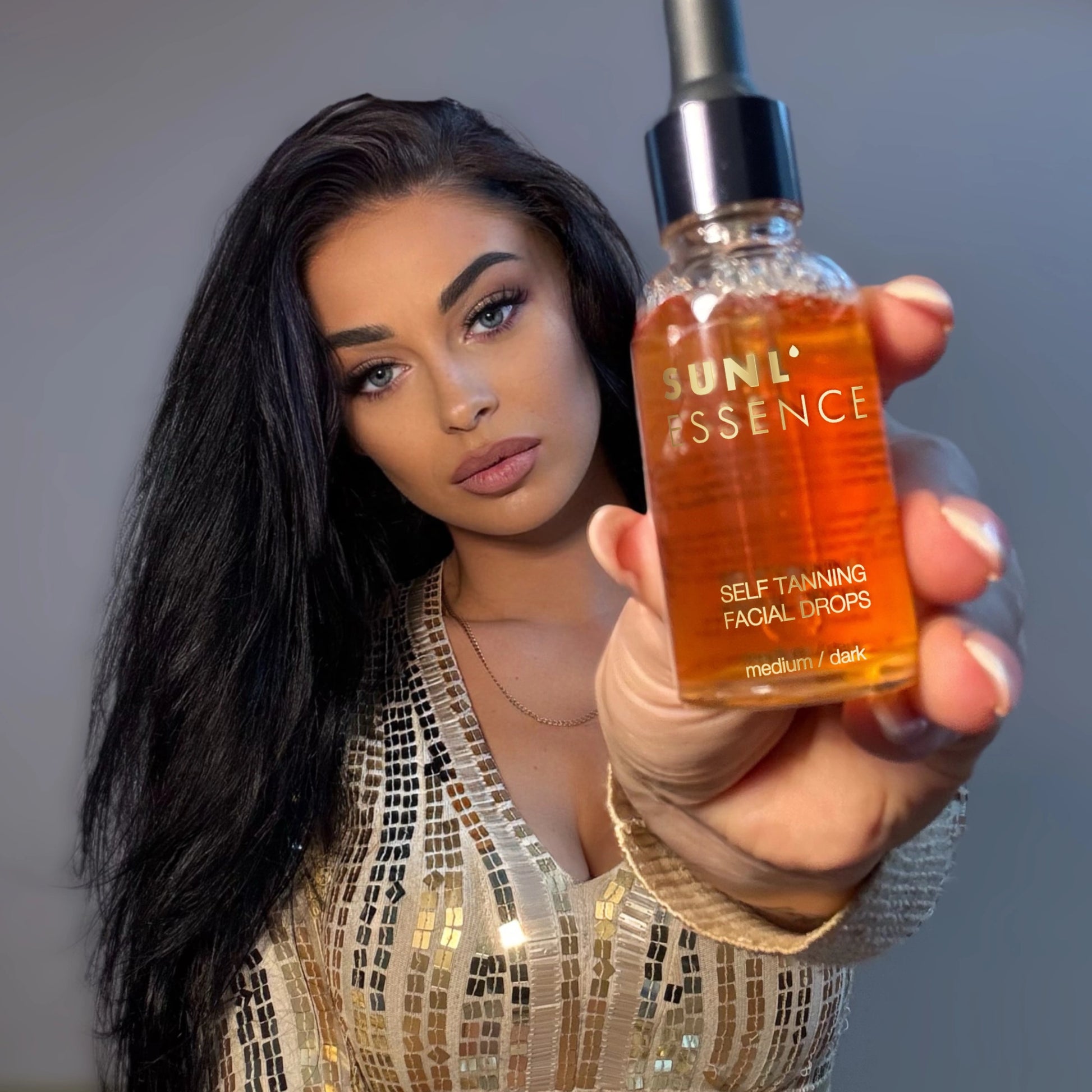 Woman with long dark hair holding a bottle of self-tanning facial drops close to the camera
