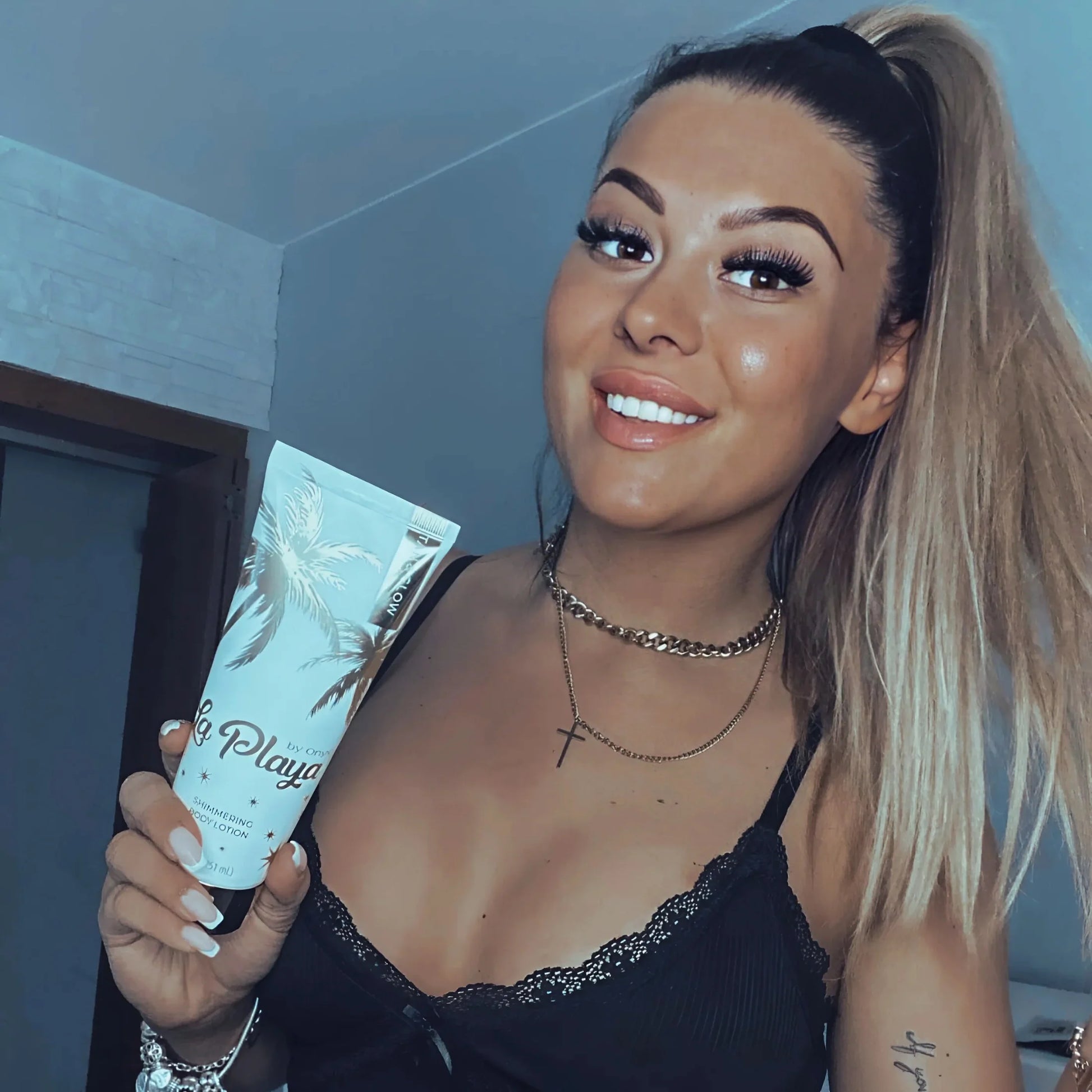 Smiling woman with blonde hair in a black top holding a tube of shimmering body cream indoors
