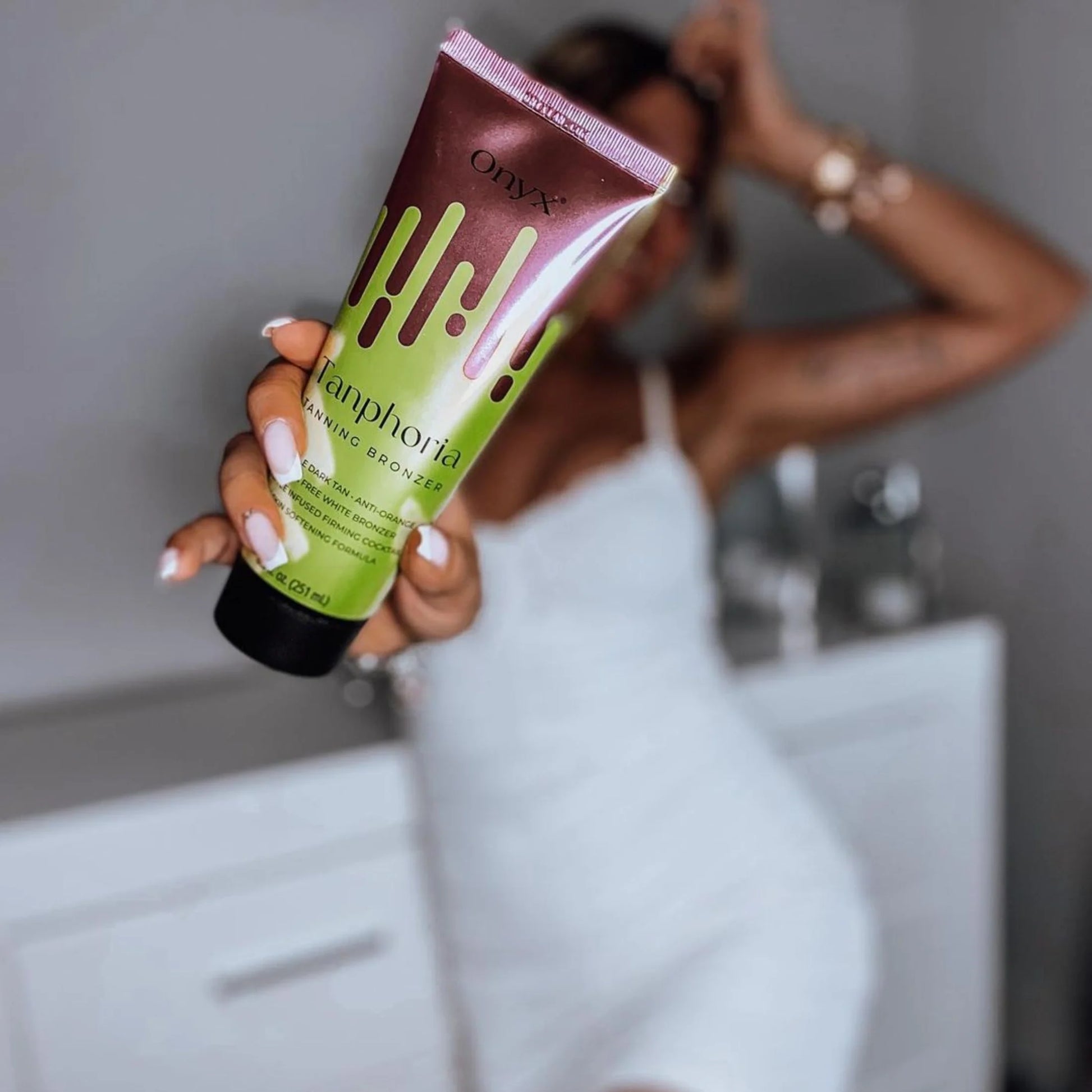 A woman in a white dress holds a Tanphoria Tanning Bronzer tube towards the camera, with the product in sharp focus and the background softly blurred.
