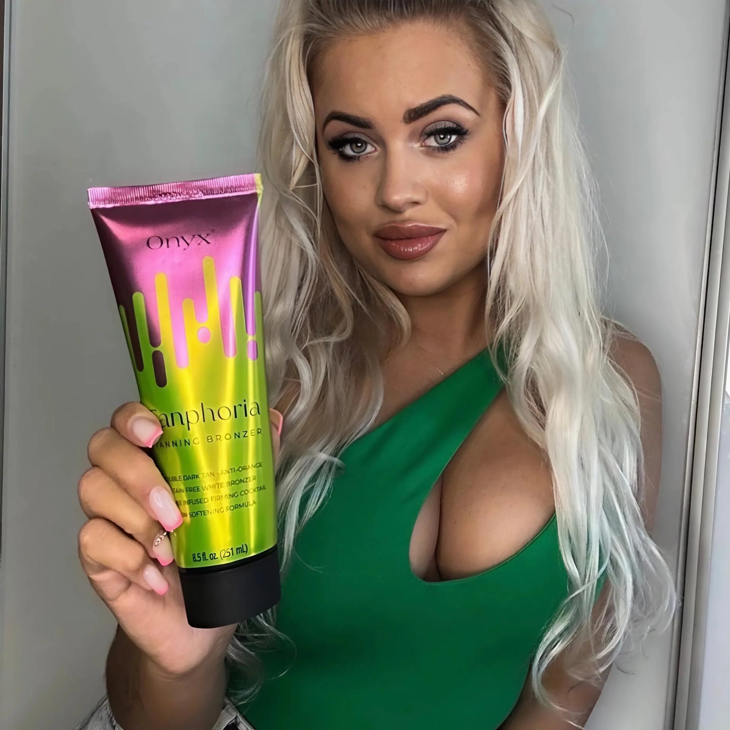 Blonde woman in a green top holds a Tanphoria Tanning Bronzer and smiles slightly, showing a sun-kissed glow and styled hair.