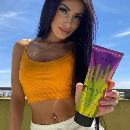 Brunette woman wearing an orange crop top holds the Tanphoria Tanning Bronzer outside on a sunny day, showing bronzed skin and relaxed confidence.
